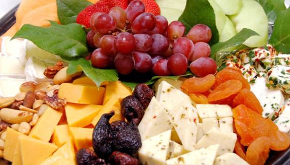 The photo showcases a beautifully arranged fruit and cheese platter from Sun Dream Yacht Charters. The platter features a variety of cheeses, including cubes of cheddar and a herbed cheese, alongside dried fruits such as figs and apricots. Fresh fruits, including a cluster of red grapes, are prominently displayed in the center, surrounded by leafy greens for garnish. The platter also includes an assortment of nuts, adding to the variety and texture of the selection. The vibrant colors and careful arrangement of the items create an inviting and elegant presentation, perfect for a luxurious yacht experience in Fort Lauderdale.