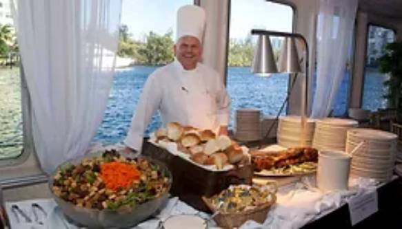 The photo shows a chef from Sun Dream Yacht Charters in Fort Lauderdale standing behind a buffet table on one of their luxurious yachts. The chef is dressed in a traditional white chef's uniform and hat, smiling at the camera. The buffet table is elegantly set up with a variety of food items, including a large bowl of salad topped with shredded carrots, a basket of bread rolls, a dish with roasted meat, and several plates and utensils. The table is covered with a white tablecloth, enhancing the upscale atmosphere. Through the windows, you can see a beautiful view of the water, adding to the picturesque setting typical of a Sun Dream Yacht Charters experience in Fort Lauderdale.